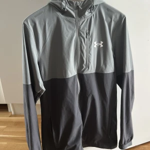 Grå vindjacka från Under Armour - Snygg tvåfärgad vindjacka från Under Armour i ljusgrått och mörkgrått. Jackan har huva, dragkedja framtill och en bröstficka med dragkedja. Perfekt för blåsiga dagar och sportiga outfits. Under Armour-logga på bröstet ger en sportig vibe.