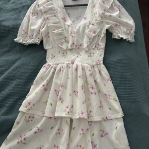 Vit blommig kortklänning Gina Tricot XS - Supersöt vit kortklänning från Gina Tricot i storlek XS. Klänningen har lila och rosa blommor, volanger längs v-ringningen och puffiga kortärmar. Smockad midja och lätt, skönt material som passar perfekt för varma dagar. Romantisk och fräsch vibe!