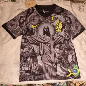 Brasil Dri-Fit fotbollströja Nike L - Unik Brasil fotbollströja från Nike med Dri-Fit material. Svart bas med detaljerat grafiskt mönster i grått och gult, inklusive Kristusstatyn och brasilianska symboler. Kortärmad med Brasil-logga och Nike Swoosh på bröstet. Perfekt för dig som vill sticka ut på planen.