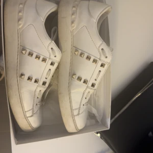 Valentino Rockstud vita sneakers - Snygga vita Valentino Rockstud sneakers i skinn med ikoniska silvriga nitar längs sidorna. Klassisk rund tå, snörning och platt sula. Perfekta för dig som vill ha en lyxig och edgy touch till din outfit.
