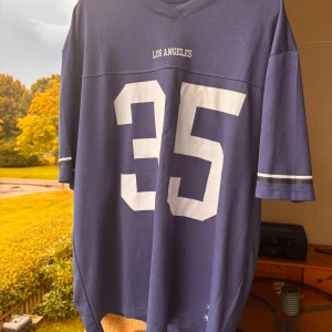 Blå Los Angeles fotbollströja M - Snygg blå fotbollströja från New Garments i storlek M, tillverkad i 100% polyester. Framsidan har stort vitt tryck med siffran 35 och texten 'Los Angeles' både fram och bak. Klassisk v-ringad hals och vita ränder på ärmarna.