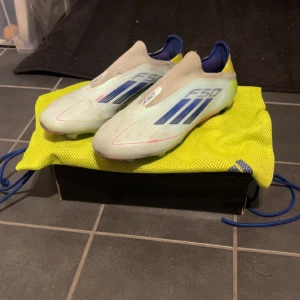 Adidas F50 fotbollsskor blå/gul - Säljer ett par Adidas F50 fotbollsskor i vitt med blå detaljer och gula samt rosa inslag på sulan. Skorna har en strömlinjeformad design utan snörning och är tillverkade i syntetmaterial. Perfekta för dig som vill sticka ut på planen. Innersulor och skopåse medföljer.