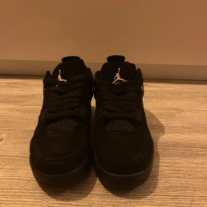 Svarta Nike Air Jordan 4 sneakers - Säljer ett par svarta Nike Air Jordan 4 sneakers med klassisk Jumpman-logga i vitt på plösen och hälen. Skorna har snörning, meshpaneler och detaljer i mocka för en clean och sportig look. Perfekta för dig som gillar streetwear och vill sticka ut.
