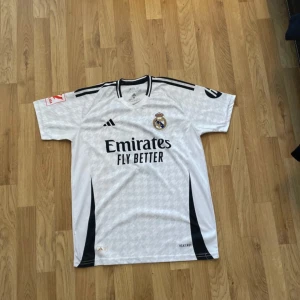 Real Madrid Rodrygo matchtröja - Säljer en vit Real Madrid fotbollströja från Adidas med Rodrygo och nummer 11 på ryggen. Tröjan har svarta detaljer, korta ärmar och ett subtilt mönster över hela tyget. Officiell klubbmärke och sponsortryck på bröstet. Perfekt för dig som älskar fotboll och Real Madrid.