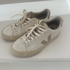 Vita sneakers från Veja med beige detaljer - Säljer ett par stilrena vita sneakers från Veja med beige V-logga på sidan. Skorna har rund tå, platt sula och snörning framtill. Tillverkade i skinn med gummisula som har hexagonmönster undertill. Perfekta för dig som gillar minimalistisk och clean stil. Skorna är måttligt använda, inköpta på Wakakuu.