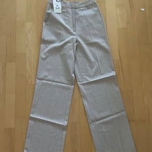 Beige randiga kostymbyxor - Snygga beige kostymbyxor med vita tunna ränder från PW Jaclin. Byxorna har raka ben, hög midja och klassisk passform. Perfekta för en clean och stilren look. Materialet är mjukt och känns bekvämt mot huden. Från BikBok 