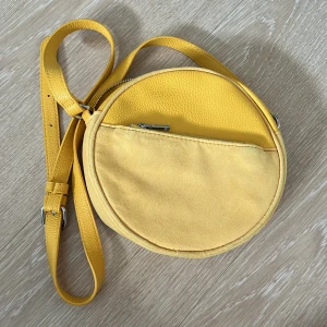 Gul rund axelväska i skinnimitation från H&M - Säljer en rund axelväska i starkt gul färg med justerbart axelband. Väskan har två fack med dragkedja, ett större huvudfack och ett mindre ytterfack. Materialet är en mix av slät och lätt strukturerad skinnimitation. Perfekt för dig som vill sticka ut med en färgklick!
