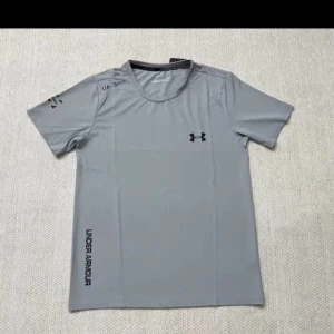 Grå träningst-shirt Under Armour - Snygg grå t-shirt från Under Armour med svart logga på bröstet och tryck på ärm och nederkant. Perfekt för träning, med korta ärmar och rund hals. Tillverkad i lätt syntetmaterial som andas och håller dig sval under passet.