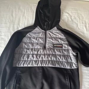 Svart och grå hoodie från McKenzie - Snygg hoodie från McKenzie i svart med grå quiltad panel framtill. Tröjan har huva med snörning, half zip och långa ärmar.