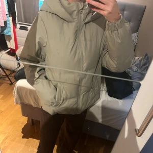 Beige pufferjacka med huva - Säljer en grön/beige pufferjacka med huva och dragkedja framtill. Jackan har en clean look, elastiska muddar vid ärmsluten och två fickor på sidorna. Perfekt för kyliga dagar och enkel att styla till olika outfits.