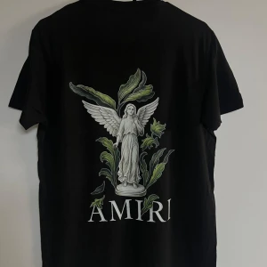 Svart AMIRI t-shirt med ängeltryck - Svart t-shirt från AMIRI med stort tryck av en ängel och gröna blad på ryggen samt AMIRI-logga. Framsidan har en stilren logga med färgstänk. Klassisk passform och korta ärmar, tillverkad i mjuk bomull. Perfekt för dig som vill sticka ut med en unik design.