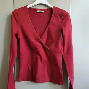 Vinröd omlottopp från Vero Moda - Vinröd långärmad omlottopp från Vero Moda med v-ringning. Topp i mjuk bomull med figurnära passform och stilren design, perfekt att styla med jeans eller kjol. Enkel men snygg detalj med omlott framtill.