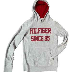 Grå hoodie Tommy Hilfiger med tryck - Snygg grå hoodie från Tommy Hilfiger med röd text 'Hilfiger Since 85' på bröstet. Tröjan har huva med rött foder och en stor magficka. Perfekt för dig som gillar sportig och avslappnad stil. Deflekten är en liten gul fläck på slutet av ena armen och en liten gul fläck i mitten, inget men ser när man bär den😁👍. Skick 7/10😁. 