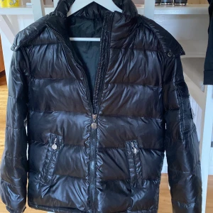 Svart pufferjacka från Moncler - Säljer en svart pufferjacka från Moncler med glansig finish och klassisk logga på ärmen. Jackan har huva, dragkedja framtill och två fickor med dragkedja. Perfekt för kalla dagar när du vill vara både varm och snygg.