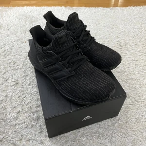 Svarta Adidas Ultraboost sneakers - Snygga helsvarta Adidas Ultraboost sneakers med stickad ovandel och klassiska tre ränder på sidan. Skorna har platt sula och snörning för perfekt passform. Perfekta för dig som gillar stilrena och bekväma sneakers.
