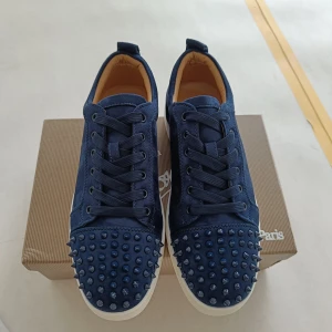 Blå mocka sneakers - Snygga sneakers i mörkblå mocka med nitar på tån och klassisk röd sula. Skorna har snörning och vit platt sula, perfekt för dig som vill sticka ut med en lyxig stil.