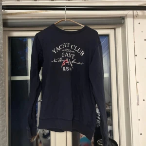 Mörkblå GANT sweatshirt Yacht Club - Mörkblå sweatshirt från GANT med Yacht Club-tryck framtill i vitt och rött. Tröjan har rund halsringning med röd detalj på insidan, långa ärmar och är tillverkad i 100% bomull. Perfekt för dig som gillar klassisk och sportig stil.
