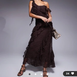 Mörkbrun spetsklänning med volanger - Säljer en lång, mörkbrun klänning i spets med volangdetaljer och oneshoulder-design. Klänningen har en romantisk look med transparenta partier och asymmetriska volanger som ger extra rörelse. Perfekt för dig som vill sticka ut med en elegant och unik stil. Klänningen har inte använts och har fortfarande prislappen på.