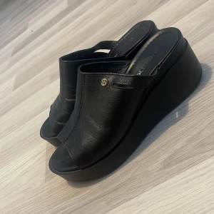 Svarta platåsandaler från Wanna - Svarta sandaler med platåsula från Wanna. Skorna har öppen tå, rund form och är tillverkade i skinn. Snygg detalj med liten metallknapp på sidan. Perfekta för dig som gillar en stilren och modern look med extra höjd.