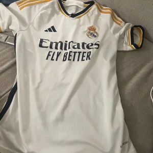 Real Madrid Bellingham matchtröja - Snygg vit Real Madrid fotbollströja från Adidas med Bellingham och nummer 5 på ryggen. Tröjan har korta ärmar, guldiga och svarta detaljer samt klubbmärke och sponsortryck framtill. Perfekt för dig som vill visa ditt stöd för laget. Pris kan diskuteras vid snabb affär 