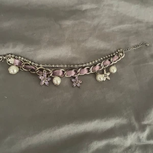Lila armband med pärlor och blommor - Snyggt armband med silverfärgad kedja och lila sidenband flätat genom länkarna. Dekorerat med hängande vita pärlor och blommor i ljuslila kristaller. Extra rad med små glittriga stenar för extra bling. Perfekt för dig som gillar söta detaljer och lite extra sparkle.