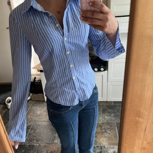 Blåvit randig skjorta från Bershka - Snygg blå skjorta med vita vertikala ränder från Bershka. Skjortan har klassisk krage, lång ärm och knäppning framtill. Figurnära passform som ger en clean och fräsch look. Perfekt att styla med jeans för en trendig vibe.