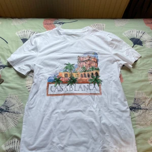 Vit t-shirt Casablanca print - Vit t-shirt från Casablanca med färgglad print på bröstet som visar en byggnad, palmer och texten 'Casablanca'. Rund hals och korta ärmar. Perfekt för dig som gillar snygga prints och vill sticka ut lite extra.
