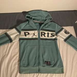 Jordan PSG hoodie i turkos och vitt  - Snygg hoodie från Jordan x PSG i turkos med vita och svarta detaljer. Stor PARIS-tryck över bröstet, PSG-logga på ärmen och klassisk huva. Dragkedja framtill och två fickor. Perfekt för dig som gillar sportig streetstyle. Det passar även till dom som har Storlek S & M