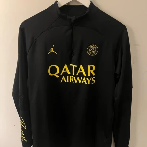  PSG långärmad tröja  - Slim fit svart fotbollströja från PSG x Jordan med gul logga och tryck. Lång ärm, halv dragkedja och broderat klubbmärke på bröstet. Materialet är mjukt och stretchigt, perfekt för träning eller match. Snygg gul Paris Saint-Germain badge och Jordan-logga.