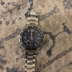 Tommy Hilfiger silverfärgad klocka - Stilren klocka från Tommy Hilfiger med svart urtavla och silverfärgat armband i metall. Urtavlan har vita detaljer, två blå subdials och tydliga siffror vid 12 och 6. Perfekt accessoar för dig som gillar klassisk design med sportig touch. Köpt för 2000kr.