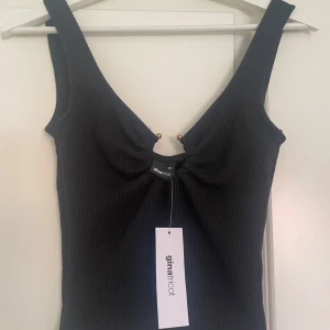 Svart ribbad linnetopp Gina Tricot - Svart ribbad linnetopp från Gina Tricot med djup v-ringning och snygg rynkdetalj framtill. Tjockare axelband och coola guldfärgade metalldetaljer vid urringningen. Perfekt att styla med jeans eller kjol.