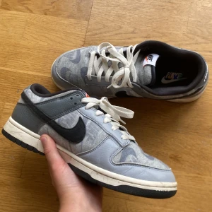 Nike Dunk Low i grått och svart - Säljer ett par Nike Dunk Low sneakers i grått och svart med vit sula och svarta detaljer. Ovandelen har ett unikt mönster med flera Nike-loggor och är tillverkad i en mix av textil och skinn. Skorna har klassisk snörning och rund tå. Perfekta för dig som gillar streetwear.