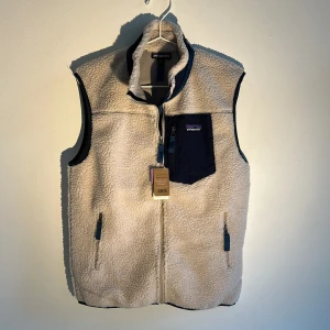 Patagonia väst - Säljer en beige fleeceväst från Patagonia med marinblå detaljer. Väldigt populär väst som passar snyggt in i många outfits.😁 Först till kvarn.
