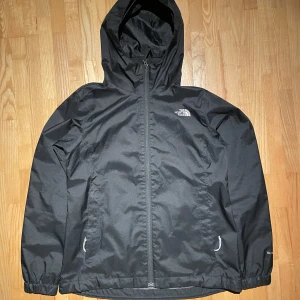 Svart vindjacka från The North Face - Svart vindjacka från The North Face med luva. Jackan har två fickor med dragkedja och elastiska muddar vid ärmsluten. Perfekt för blåsiga dagar och enkel att matcha med allt. Snygg och funktionell design.