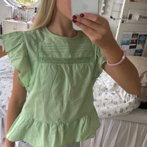 Grön blus med volang från H&M Divided - Supersöt ljusgrön blus från H&M Divided med volangkant nertill och volangärmar. Stängs med knapp i nacken.