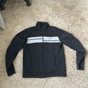 Svart vindjacka från BOSS med vit rand - Svart vindjacka från BOSS med två vita horisontella ränder över bröstet och diskret BOSS-logga. Jackan har hel dragkedja framtill, ståkrage och ribbade muddar vid ärmslut och nederkant. Perfekt för en clean och sportig look.