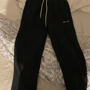 Svarta mjukisbyxor från Champion - Svarta mjukisbyxor från Champion med vit snörning i midjan och broderad logga på ena benet. Byxorna har en avslappnad passform och är perfekta för chill dagar. Tillverkade i mjukt material som känns skönt mot huden.