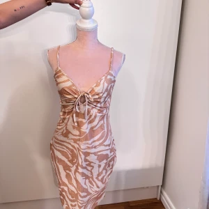 Beige och vit zebra kortklänning - Supersnygg kort klänning med beige och vitt zebramönster. Smala axelband, v-ringning och knytning framtill ger en trendig look. Materialet är ribbat och klänningen sitter figurnära, perfekt för dig som vill sticka ut.