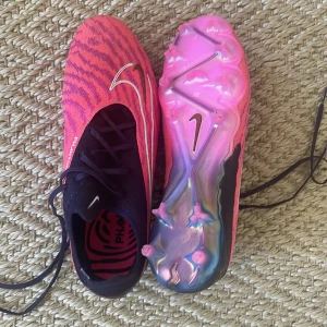 Nike Phantom rosa fotbollsskor - Skorna är använda en gång, men var lite för stora för mig på bredden då jag har väldigt smala fötter. Nike Phantom fotbollsskor i en riktigt fet rosa färg med lila zebramönster och svarta detaljer. Skorna har snörning, vit Nike swoosh och en glansig rosa sula med dobbar. Ovandelen är i syntetmaterial och designen är riktigt sportig och modern.