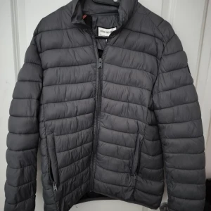 Mörkgrå pufferjacka Shine Original - Säljer en mörkgrå pufferjacka från Shine Original med horisontella sömmar, hög krage och dragkedja framtill. Jackan har två fickor med dragkedja och en snygg patch på ärmen. Perfekt för kyliga dagar och enkel att matcha med det mesta.