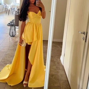 Gul långklänning med snörning - Supersnygg gul långklänning med omlott-design och snörning i ryggen. Klänningen har axelbandslös överdel med markerad byst och volangdetaljer framtill. Materialet är glansigt och känns lyxigt, perfekt för bal eller student. Passformen är normal och kjolen har en cool slits.