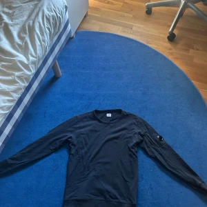 Svart sweatshirt från C.P. Company - Svart långärmad sweatshirt från C.P. Company med klassisk rund hals och ikonisk linsdetalj på ärmen. Tröjan har en liten ficka med dragkedja på vänster ärm och är tillverkad i mjuk bomull. Perfekt för en clean och stilren look. Pris går att diskuteras 