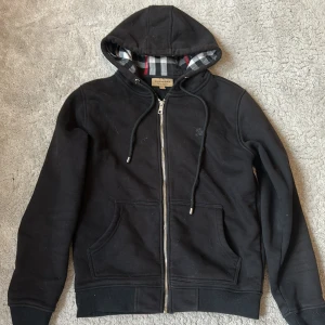 Svart hoodie från Burberry med dragkedja - Svart hoodie från Burberry med klassisk huva, dragkedja framtill och snörning. Insidan av huvan har Burberrys rutiga mönster. Tröjan har två fickor framtill och är tillverkad i mjukt material, perfekt för kyliga dagar.