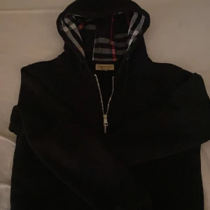 Svart Burberry hoodie med dragkedja - Svart hoodie från Burberry med klassiskt rutigt foder i huvan och broderad logga på bröstet. Tröjan har dragkedja framtill, snörning i huvan och långa ärmar. Perfekt för dig som gillar stilrena och exklusiva plagg med en twist. Har använt några gånger innan.