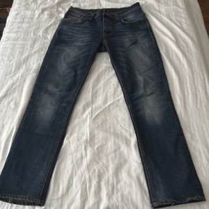 Mörkblå jeans från Nudie Jeans - Säljer ett par mörkblå jeans från Nudie Jeans med klassisk femficksdesign och snygga slitningar. Jeansen har orange kontrastsömmar och Nudie-logga på bakfickan samt läderpatch i midjan. Perfekta för dig som gillar en tidlös och avslappnad stil. W: 30 L: 32 