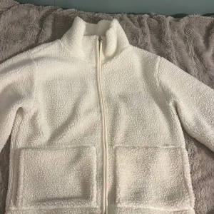 Vit teddyjacka med dragkedja - Mysig vit teddyjacka med hög krage och två stora fickor framtill. Jackan har hel dragkedja och är tillverkad i fluffigt fleece-material som håller dig varm. Perfekt för kyliga dagar och enkel att styla med det mesta. Långärmad och i en klassisk, rak passform.