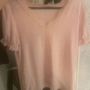 Söt rosa blus med spetsdetaljer - Supersöt ljusrosa blus med broderade hålmönster och spets längs v-ringningen. Puffiga korta ärmar med volangkant ger en romantisk vibe. Perfekt för dig som gillar en feminin och gullig stil.