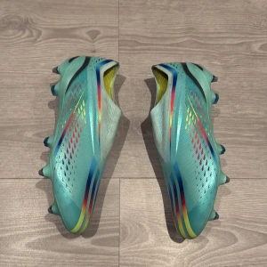 Adidas X Speedflow fotbollsskor turkos - Säljer ett par Adidas X Speedflow fotbollsskor i en fräsch turkos färg med regnbågsfärgade detaljer och gula insidor. Skorna har en strömlinjeformad design och är utan snöre, metall dobbar. Perfekta för dig som vill sticka ut på matchen!