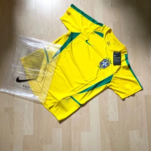 Brasilien retro - Brazil Nike Fotbollströja | CBF Officiell Tröja  Officiell brasiliansk landslags fotbollströja från Nike. Klassisk gul design med gröna detaljer och broderat CBF-emblem. Perfekt för samlare, fotbollsfans, eller att bära under matcher. Skick: Ny med etiketter. Storlek: M  Ett fantastiskt föremål för alla fotbollströjesamlingar eller ett måste för brasilianska supportrar!  Brasilien fotbollströja, Nike Brasilien tröja, CBF officiell kit, Brasilien fotbollströja, VM-tröja, vintage fotbollströja.