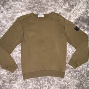 Olivgrön sweatshirt från Stone Island - Säljer en olivgrön sweatshirt från Stone Island Junior med klassisk logga på ärmen. Tröjan har rund hals, långa ärmar och ribbade muddar. Perfekt för dig som gillar streetwear och vill ha något stilrent men ändå med attityd. Kom privat för mer info!💯